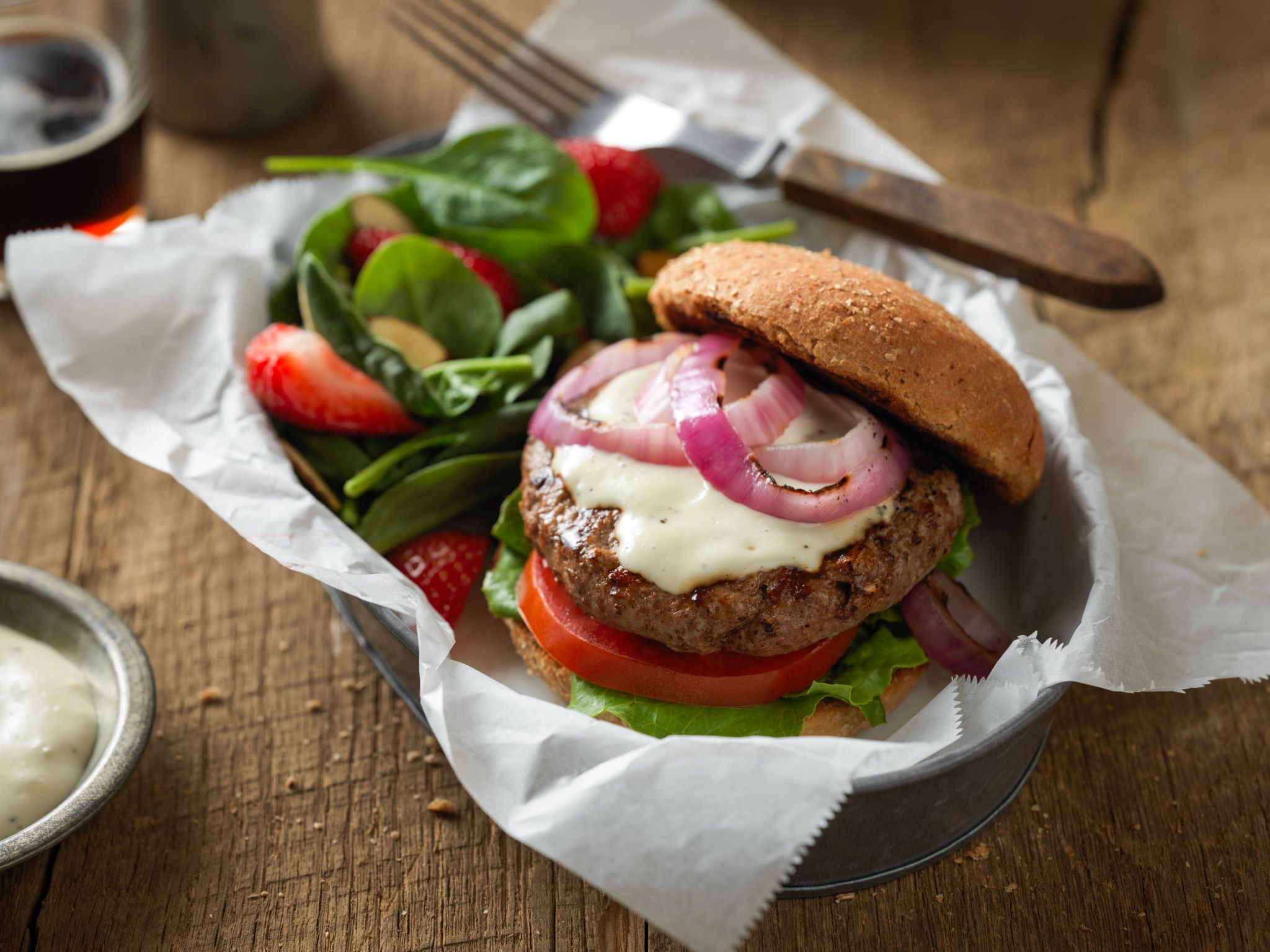 Bear River Valley Beef - Bulk Beef Juicy beef burger topped with melted cheese, red onions, and fresh lettuce, served on a whole grain bun alongside a salad of spinach and strawberries, with a small bowl of dressing on a rustic wooden table.