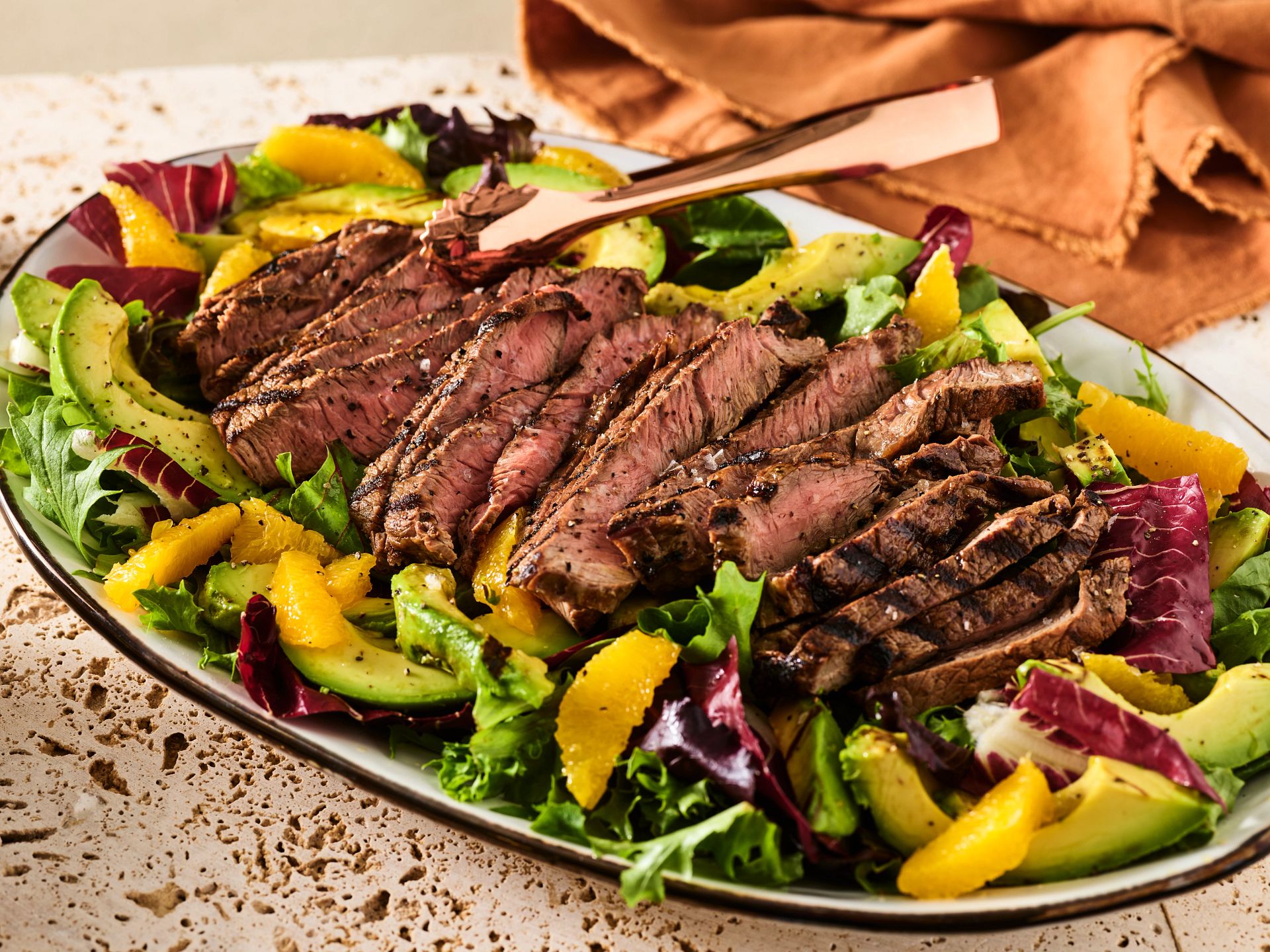Bear River Valley Beef - ranch package Grilled steak slices served on a bed of mixed greens, avocado, and orange segments, arranged on a decorative platter with serving utensils nearby.