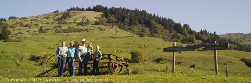 Liz Klopper - 20100508-Klopper Family-0023 Family standing in a green pasture with a vintage farm implement, surrounded by rolling hills and trees, showcasing rural life and farming heritage.