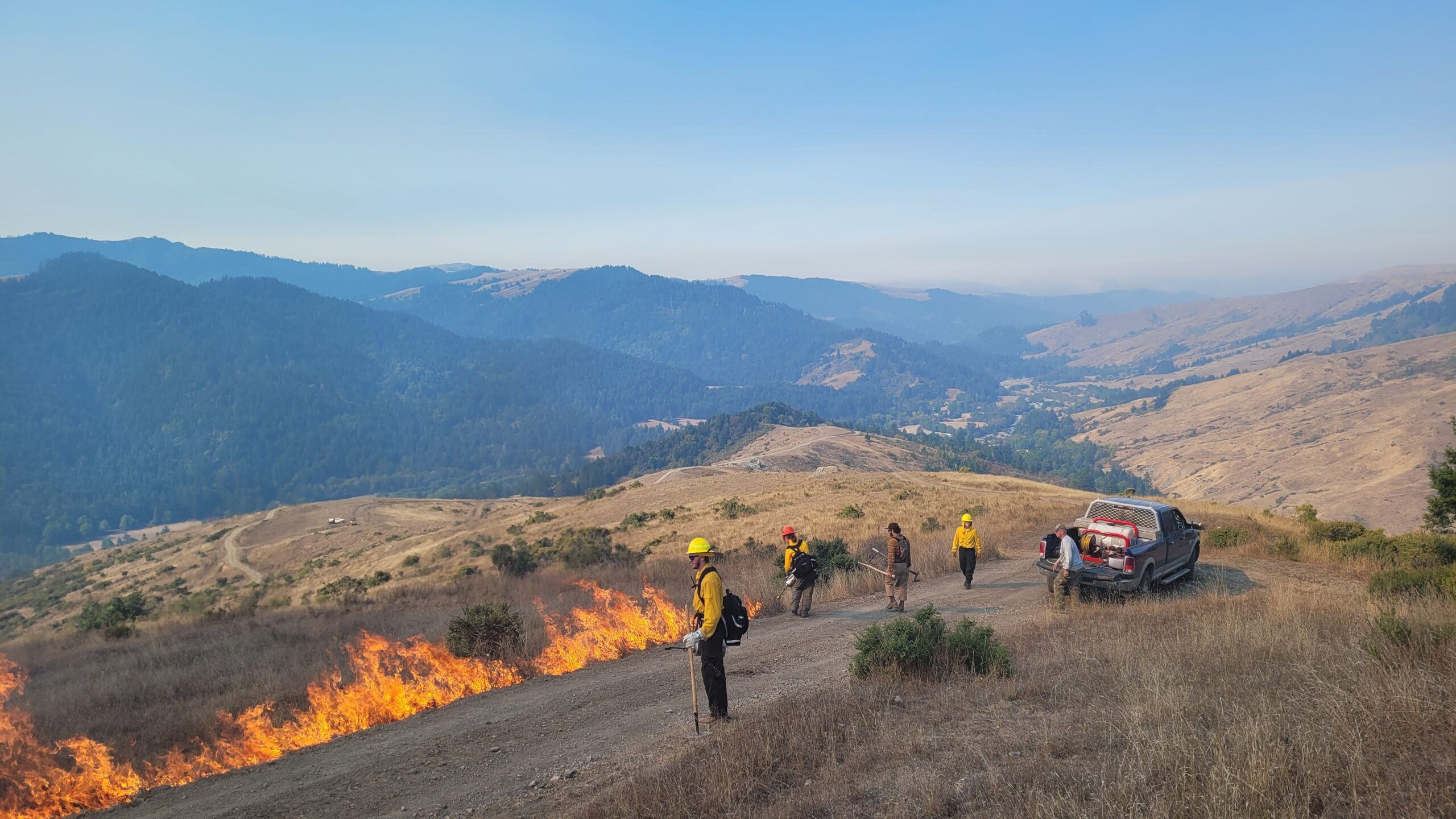 Liz Klopper - 20230922_105009 Firefighters conducting controlled burn in a mountainous area, with flames visible along a dirt road, surrounded by dry grass and hills. A truck is parked nearby as team members work to manage the fire.