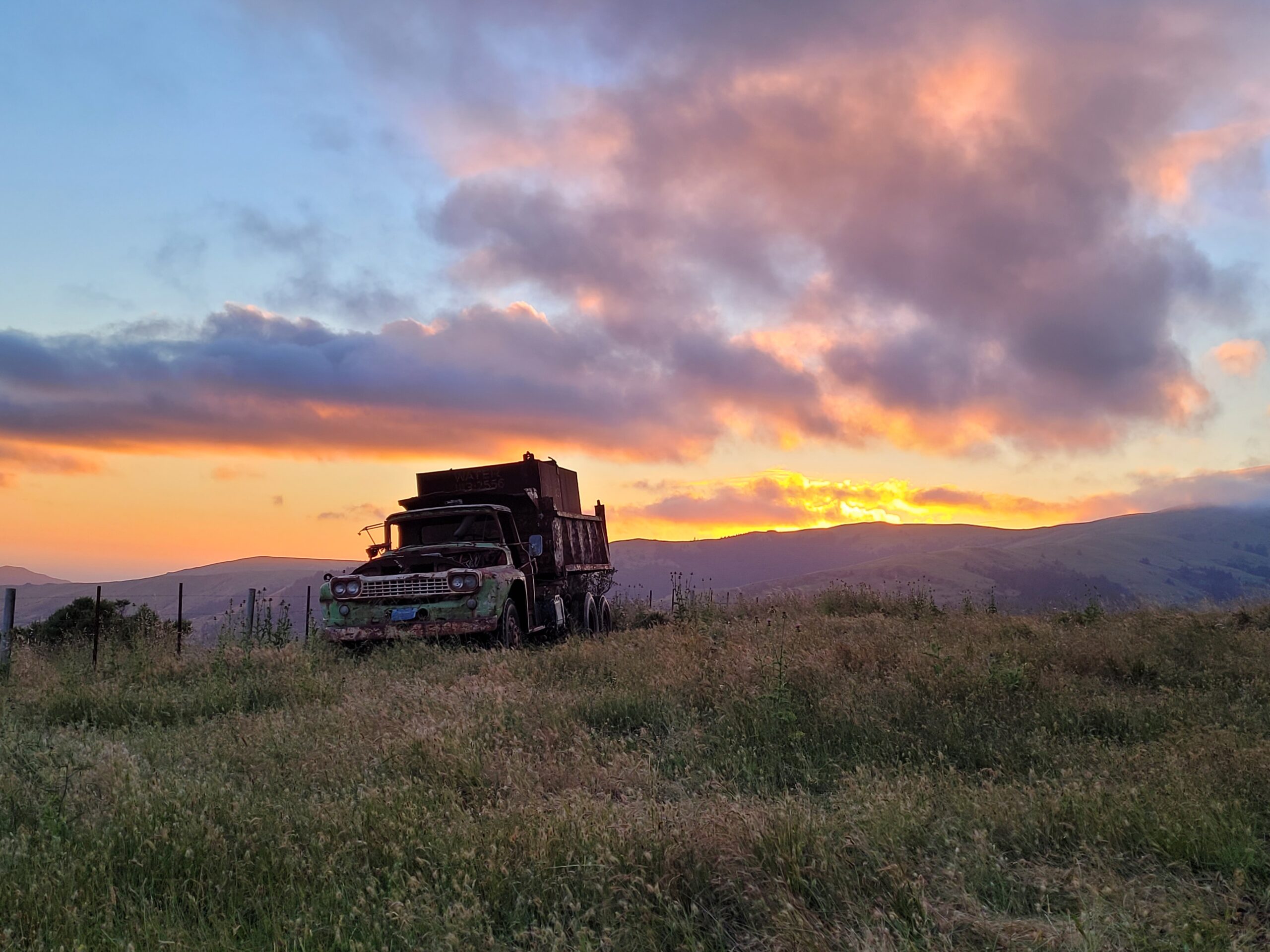 Liz Klopper - old dump truck Abandoned vintage truck in a grassy field at sunset, with colorful clouds and rolling hills in the background, showcasing the beauty of rural landscapes.